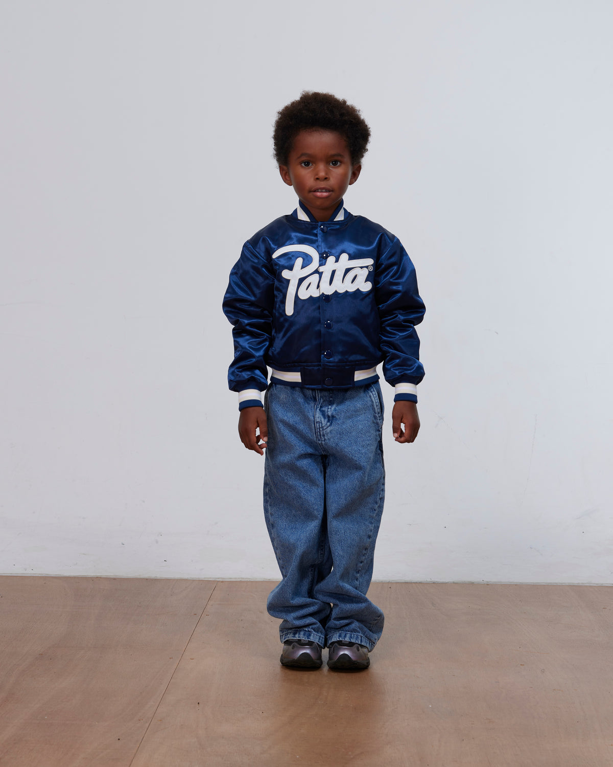 Boy wearing the Patta Kids Satin Bomber Jacket in Black Iris paired with denim jeans in a studio.