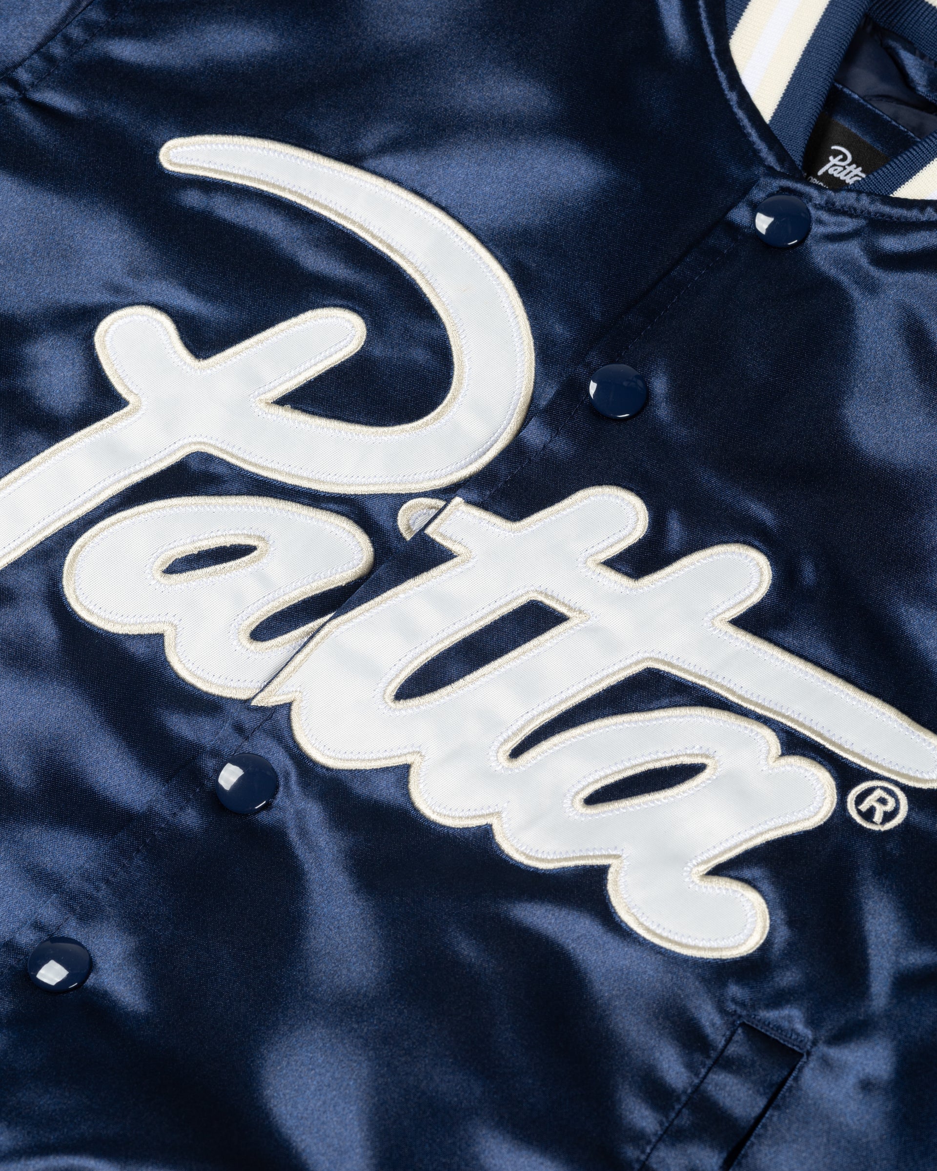 Detail of the white script logo appliqué on the Black Iris Patta kids satin bomber jacket.